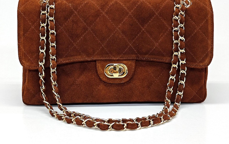 May include: A brown suede handbag featuring a quilted diamond pattern, a gold-tone metal turn-lock closure, and a long chain strap interwoven with matching brown suede material. The rectangular bag has a structured flap and polished gold hardware.