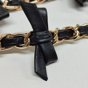 K&ouml;nnte beinhalten: Nahaufnahme einer schwarzen Lederkette mit goldfarbenen Gliedern. Die Kette ist mit kleinen schwarzen Lederschleifen verziert. Das Design ist elegant und detailliert, mit Fokus auf Textur und Kontrast der Materialien.