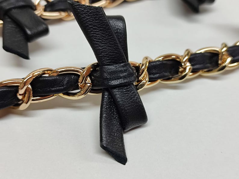 K&ouml;nnte beinhalten: Nahaufnahme einer schwarzen Lederkette mit goldfarbenen Gliedern. Die Kette ist mit kleinen schwarzen Lederschleifen verziert. Das Design ist elegant und detailliert, mit Fokus auf Textur und Kontrast der Materialien.