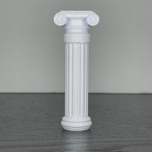 May include: A white, miniature Ionic column with a fluted shaft and decorative scrollwork at the capital. The column stands upright against a neutral gray background, showcasing its classical architectural design. The column is a decorative item.