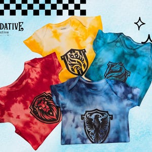 May include: Four short-sleeve baby bodysuits in tie-dye patterns of yellow, red, teal, and blue. Each garment features a black shield-shaped graphic on the chest depicting a lion, an owl, a dragon, or a phoenix. The text Alnerdative Collective is visible.