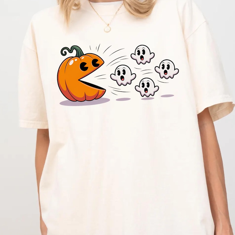 Puede incluir: Camiseta de color crema con un dise&ntilde;o de calabaza de dibujos animados persiguiendo fantasmas. La calabaza es naranja con un tallo verde y una boca muy abierta. Los fantasmas son blancos con ojos y bocas negras. Tema de Halloween juguet&oacute;n.