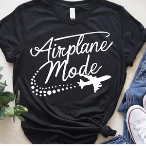 May include: Black t-shirt with the words "Airplane Mode" in a white script font. A white airplane graphic and dotted trail are below the text. The shirt is styled with jeans and white sneakers.