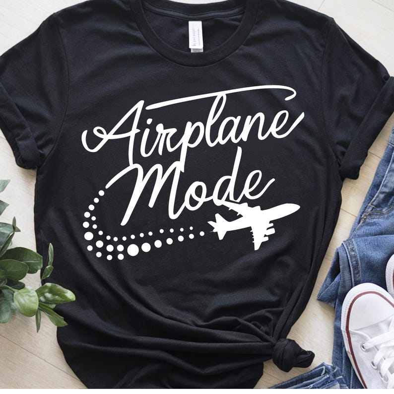 May include: Black t-shirt with the words "Airplane Mode" in a white script font. A white airplane graphic and dotted trail are below the text. The shirt is styled with jeans and white sneakers.