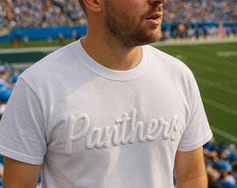 May include: A white t-shirt with the word "Panthers" in a raised, textured font. The person is standing in a stadium with a green field and blue seating in the background. The shirt is short-sleeved.