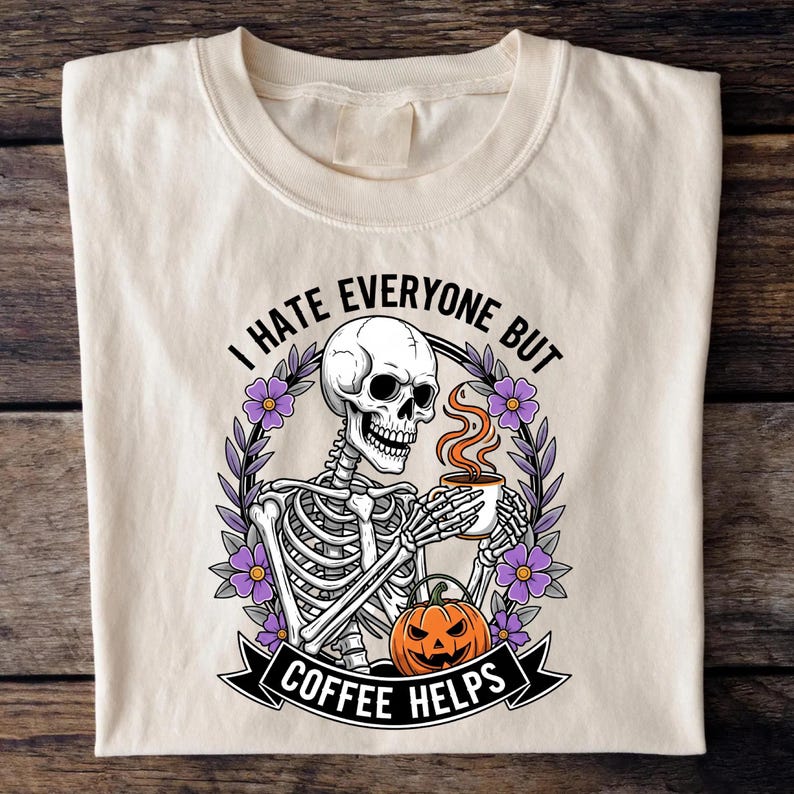 Puede incluir: Camiseta color crema con un gr&aacute;fico de un esqueleto sosteniendo una taza de caf&eacute; y una calabaza de Halloween. El texto dice "I HATE EVERYONE BUT COFFEE HELPS" en negro. El dise&ntilde;o incluye flores moradas y una pancarta negra.