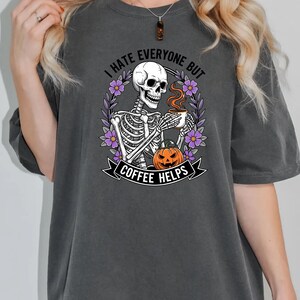 Puede incluir: Camiseta gris oscuro con un esqueleto sosteniendo una taza de caf&eacute;, rodeado de flores. El texto dice "I HATE EVERYONE BUT COFFEE HELPS" con una calabaza. Un dise&ntilde;o humor&iacute;stico para los amantes del caf&eacute;.