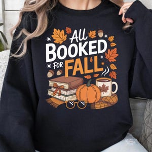 May include: Black sweatshirt with the text "All Booked for Fall" in white and orange. The design includes fall-themed illustrations of leaves, acorns, books, a pumpkin, a mug of coffee, and sunglasses.
