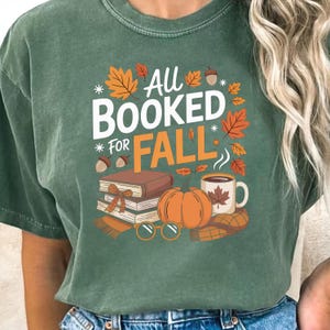 May include: Green t-shirt with the text "All Booked For Fall" in white and orange. The design includes fall-themed illustrations such as leaves, a pumpkin, books, and a coffee mug.