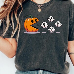 Puede incluir: Camiseta gris oscuro con un dise&ntilde;o de Halloween. Una calabaza naranja con una boca al estilo Pac-Man persigue a cuatro fantasmas blancos. El dise&ntilde;o est&aacute; en una camiseta gris oscuro de manga corta.