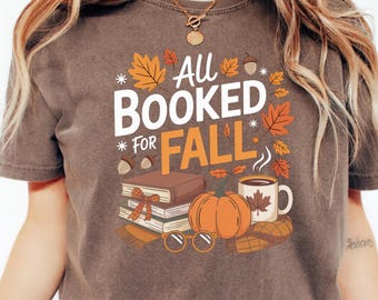 May include: Brown t-shirt with the text "All Booked for Fall" in white and orange. The graphic includes a stack of books, a pumpkin, a mug of coffee, and fall leaves. A cozy autumn-themed design.