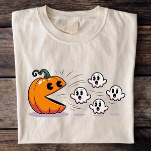 Puede incluir: Camiseta color crema con un gr&aacute;fico tem&aacute;tico de Halloween. El dise&ntilde;o incluye una calabaza naranja con una boca tipo Pac-Man persiguiendo a cuatro fantasmas blancos. La calabaza tiene un tallo verde y los fantasmas tienen expresiones de sorpresa.