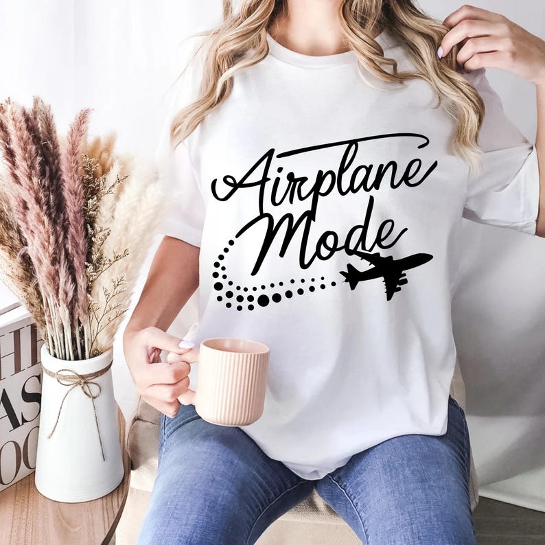 May include: White t-shirt with the words "Airplane Mode" in a stylish script, accompanied by a dotted trail and a silhouette of an airplane. The shirt is worn with blue jeans and a pink mug.
