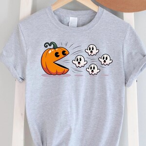 Puede incluir: Camiseta gris claro con un dise&ntilde;o tem&aacute;tico de Halloween. El dise&ntilde;o muestra una calabaza naranja con una boca al estilo Pac-Man persiguiendo a cuatro fantasmas blancos. La camiseta est&aacute; colgada en una percha de madera.