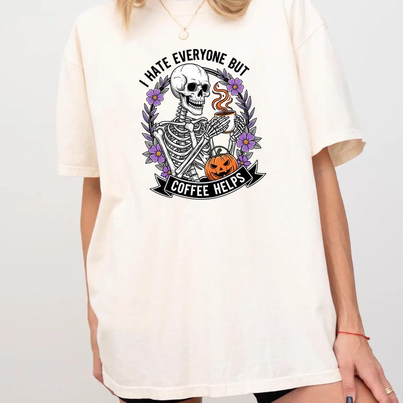 Puede incluir: Camiseta color crema con un esqueleto sosteniendo una taza de caf&eacute;, rodeado de flores moradas y una calabaza. El texto dice "I HATE EVERYONE BUT COFFEE HELPS". Una camiseta gr&aacute;fica con tem&aacute;tica de Halloween.