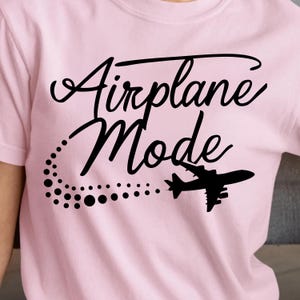 May include: A light pink t-shirt with the words "Airplane Mode" in a black script font. A black airplane silhouette is featured below the text, with a dotted trail. The shirt is a crew neck.