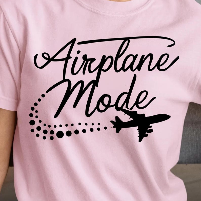May include: A light pink t-shirt with the words "Airplane Mode" in a black script font. A black airplane silhouette is featured below the text, with a dotted trail. The shirt is a crew neck.