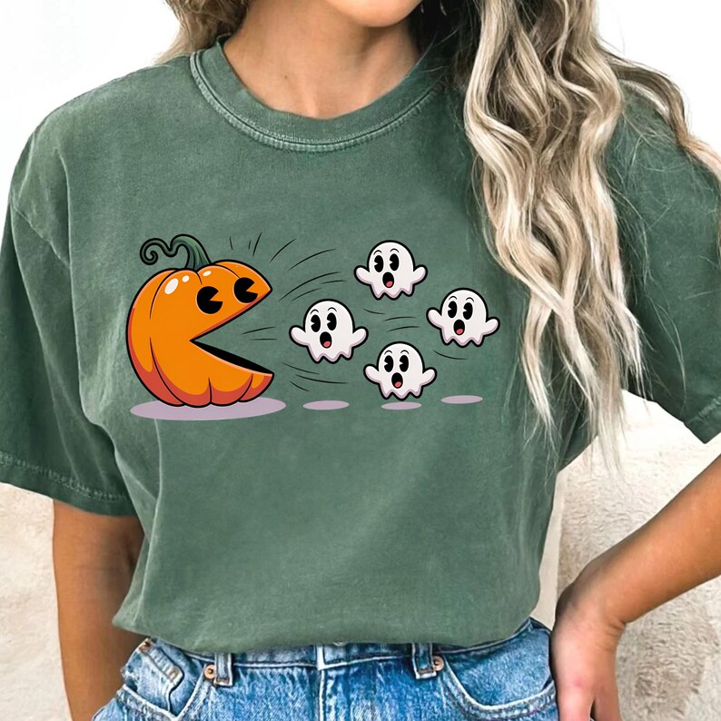 Puede incluir: Camiseta verde con un dise&ntilde;o de Halloween. El dise&ntilde;o muestra una calabaza naranja con una boca al estilo Pac-Man persiguiendo a cinco fantasmas blancos. La camiseta tiene mangas cortas y cuello redondo.