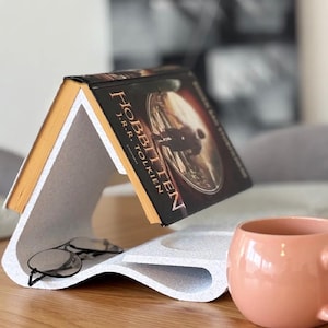 Pode incluir: Um suporte de livro branco com um design curvo, segurando uma cópia de "O Hobbit" de J.R.R. Tolkien. Um par de óculos repousa sobre o suporte, e uma caneca rosa está por perto. O suporte do livro está sobre uma superfície de madeira.