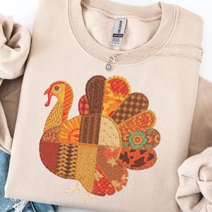 Könnte beinhalten: Beiges Sweatshirt mit Rundhalsausschnitt und einem bunten Patchwork-Truthahn-Design. Der Truthahn hat einen roten Kopf, einen orangefarbenen und gelben Körper und einen Fächer aus gemusterten Federn in Braun-, Orange- und Gelbtönen. Eine silberne Halskette mit Sonnenanhänger ist ebenfalls sichtbar.