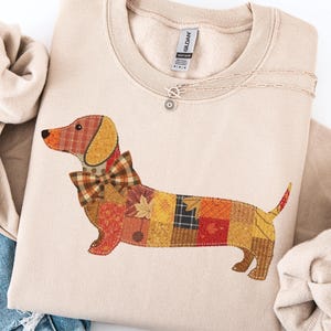 May include: Beige sweatshirt with a patchwork dachshund design in autumnal colours. The dog has a plaid bow tie and is surrounded by folded fabric and a silver necklace. The sweatshirt is a Gildan brand.