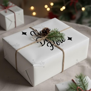 May include: A white gift box wrapped in paper, tied with twine, and adorned with a pinecone and evergreen sprigs. The words "Open Here" are printed on the front. Additional gift boxes are in the background.