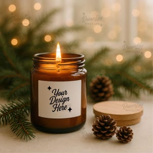 Op de afbeelding: Een brandende bruine glazen kaarsenpot met een wit etiket met de tekst "Your Design Here". De kaars is omgeven door dennennaalden, dennenappels en een houten deksel. De achtergrond heeft vage lichten.