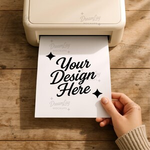 May include: A white sheet of paper with the text "Your Design Here" in black script is being printed from a cream-colored printer. The paper is held by a person with a beige sweater on a wooden surface.