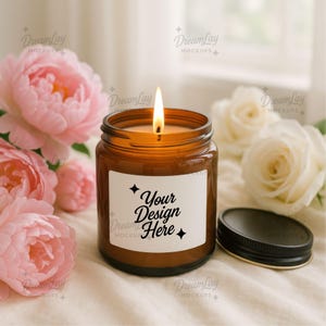 May include: A lit amber glass candle jar with a black lid, and a white label that says "Your Design Here". The candle is surrounded by pink peonies and white roses, creating a soft, floral ambiance.