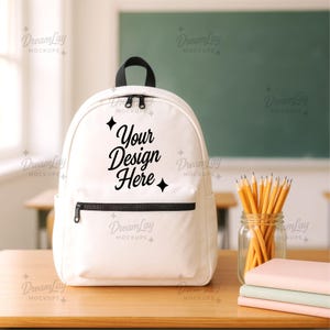 May include: A white backpack with a black zipper and handle, featuring the text "Your Design Here." The backpack is on a wooden desk, with pencils in a jar and books nearby. The background includes a chalkboard and school desks.