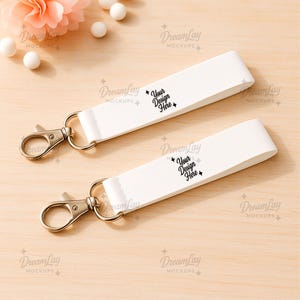 Peut inclure: Deux porte-clés blancs avec des fermoirs et des anneaux argentés. Chaque porte-clés porte l'inscription "Your Design Here" en noir. Les porte-clés sont présentés sur une surface en bois clair, avec des éléments décoratifs en arrière-plan.