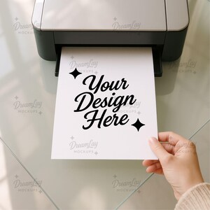 May include: A white sheet of paper with the text "Your Design Here" is being printed from a printer. The paper is held by a person's hand. The printer is gray and black.