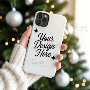 以下が含まれることがあります： スタイリッシュな筆記体で「Your Design Here」と書かれた白い携帯電話ケース。 カメラの切り抜きがあるケースは、銀色のオーナメントとライトが飾られたクリスマスツリーのぼやけた背景の前に保持されています。