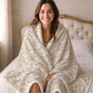 May include: A cream-coloured blanket featuring a black dandelion pattern. The blanket has a soft, fluffy white trim. The blanket is draped over a person sitting on a bed. A cosy home decor item.