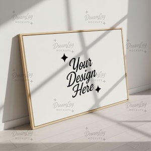 May include: A rectangular canvas with a light wood frame, leaning against a white wall. The canvas displays the text "Your Design Here" in a stylish black script. The scene is illuminated by natural light, creating shadows.