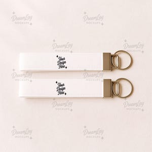 May include: Two white keychains with silver-colored hardware and key rings. Each keychain has the text "Your Design Here" printed in black. The keychains are displayed on a light pink surface.