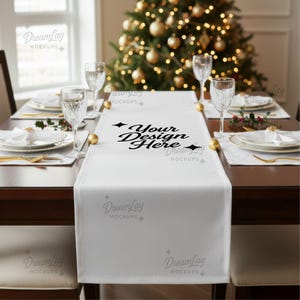Op de afbeelding: Een witte tafelloper met de tekst "Your Design Here" ligt op een donkere houten tafel. De tafel is gedekt voor een feestelijke maaltijd met gouden accenten, witte borden en kristallen glazen. Op de achtergrond staat een kerstboom.