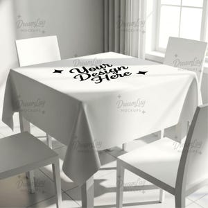 May include: A white square tablecloth with the text "Your Design Here" in black script is draped over a white table. Four white chairs surround the table, creating a clean, minimalist aesthetic. The scene is well-lit, suggesting a bright interior.