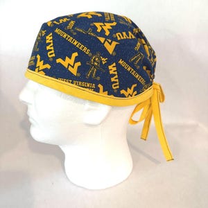 May include: A navy blue scrub cap featuring a yellow West Virginia University Mountaineers print with the text WVU and Mountaineers. The cap has a solid yellow trim along the bottom edge and includes two yellow fabric ties at the back for adjustment.