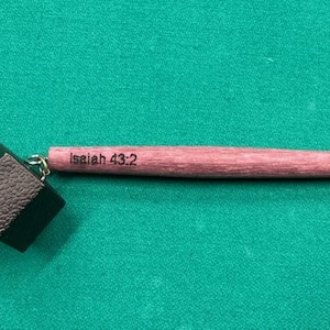 May include: A keychain with a dark green square and a cylindrical wooden piece. The wooden piece is light red and has the text "Isaiah 43:2" engraved on it. The keychain is on a green surface.