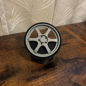 Puede incluir: Una rueda decorativa gris y negra, con un diseño de cinco radios. La rueda está colocada sobre una base negra, y la estética general sugiere una pieza de coche en miniatura o un modelo. El artículo se muestra sobre una superficie de madera.