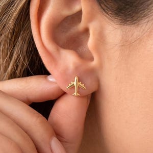 Può includere: Primo piano di un orecchino a bottone a forma di aereo color oro. L'orecchino è una piccola e dettagliata replica di un aereo, con ali, fusoliera e coda. L'orecchino è indossato su un orecchio.