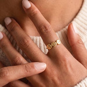 May include: A gold-toned ring featuring three small, circular charms attached to a thin band. The centre charm displays an embossed heart symbol, while the two flanking charms are each engraved with a single uppercase letter, specifically W and H.