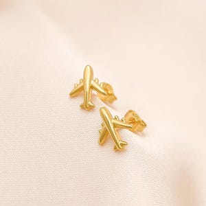 以下が含まれることがあります： 金色の飛行機スタッドピアス2つ。ピアスは小さく、詳細で、柔らかい薄いピンク色の布の背景に配置されています。飛行機のデザインは、翼、胴体、尾を備えています。