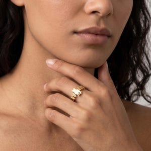 May include: A gold-toned ring featuring a central cat silhouette motif flanked by the letters M and M. The band is thin and metallic, designed to sit on a finger, showcasing a polished finish that reflects light across its smooth, curved surface.