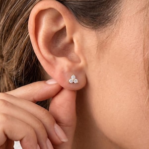 May include: A close-up of a gold stud earring with three small, round, clear gemstones. The earring is set on a person's ear. The person's hand is touching the ear.