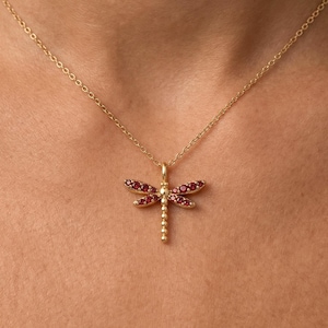 Puede incluir: Un collar de tono dorado con un colgante de libélula. La libélula tiene cuatro alas incrustadas con pequeñas piedras rojas vibrantes y una cola segmentada con cuentas. El colgante cuelga de una cadena fina y delicada de tono dorado.