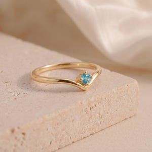 Può includere: Un anello in oro a forma di V con una singola pietra preziosa rotonda di colore blu brillante incastonata in una montatura a quattro griffe. La fascia ha una finitura liscia e lucida e segue un contorno a chevron, mettendo in risalto la pietra.