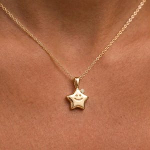 Könnte beinhalten: Eine goldfarbene Halskette mit einem sternförmigen Anhänger. Der Anhänger hat ein lächelndes Gesicht und einen kleinen Diamanten. Die Halskette wird um den Hals getragen.