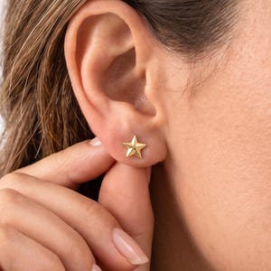 May include: A gold-colored star-shaped stud earring is worn on an ear. The earring is a five-pointed star with a slightly textured surface. The earring is being touched by a person's fingers.